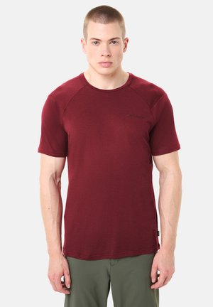 Giovane uomo con capelli corti che indossa una maglietta a maniche corte color vinaccia e pantaloni verdi, in piedi contro uno sfondo bianco.