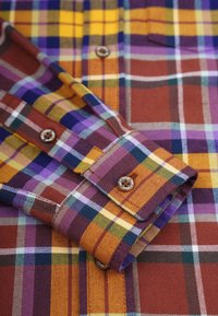 Chemise en plaid avec des tons de brun, jaune, violet et vert. Tissu en coton, poignets à boutons et boutons contrastés. Col et poche visibles.