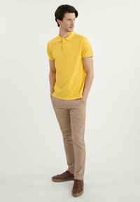 Żółta koszulka polo z szarymi akcentami, krótkie rękawy i plisą na trzy guziki, połączona z brązowymi spodniami i brązowymi trampkami.