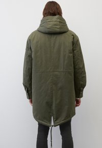 Olivgrön, lång parka med huva, med dragsko i nederkant och knäppte ärmslut. Slät tyg med en något matt yta.