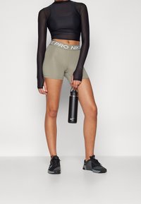 Czarny długi rękaw, top croptop, oliwkowe szorty z pasem Nike Pro, trzymający czarną butelkę na wodę, w czarnych butach sportowych.