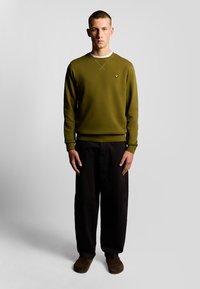 Felpa verde oliva con scollo tondo e polsini a coste, abbinata a pantaloni neri larghi e scarpe slip-on marroni. Accentuata con un logo.