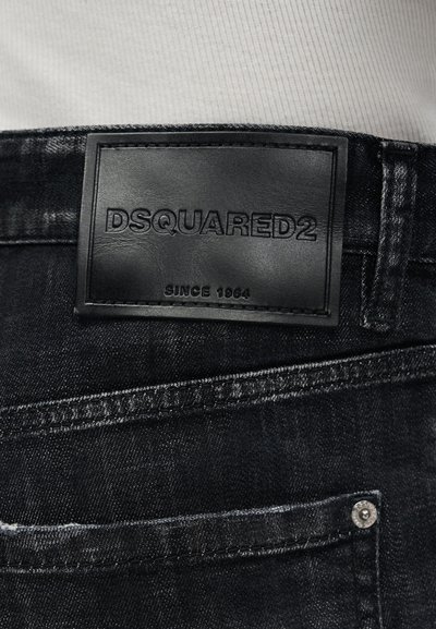 Μαύρο δερμάτινο ταμπελάκι με ανάγλυφο το "DSQUARED2 SINCE 1964" στη ζώνη από σκούρο τζιν, φορεμένο με λευκό ύφασμα από πάνω.