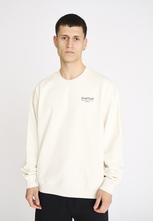 Ung mand med kort mørkt hår iført en cremefarvet sweatshirt med teksten "Social Souls" på brystet, stående foran en ensartet hvid baggrund.