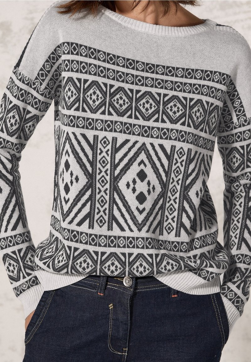 Femme portant un pull gris avec des motifs géométriques en forme de losanges noirs et un jean bleu foncé, les mains dans les poches, vue du cou jusqu’aux hanches.