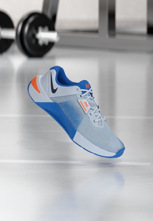 Chaussure de sport gris clair et bleu avec des accents orange, logo Nike et design moderne de la semelle sur un sol de gym brillant près des poids.