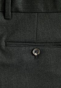 Tejido negro texturizado con un bolsillo plano y un detalle de botón de cuatro agujeros. El material es tejido y presenta un patrón fino.