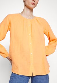 Camisa laranja de botão, com mangas longas, tecido texturizado, decote franzido e corte reto, usada sobre jeans azuis.