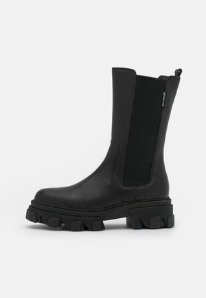 Botte mi-mollet en cuir noir avec panneaux latéraux élastiques, semelle crantée épaisse et languette au dos, vue de profil.