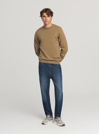 Beżowy sweter dziany z okrągłym dekoltem, zestawiony z ciemnoniebieskimi dżinsami. Model nosi szare buty sportowe, stoi na tle jednolitego tła.