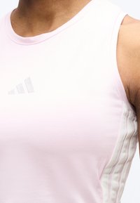 Κοντινό πλάνο ενός ατόμου που φοράει αμάνικο αθλητικό μπλουζάκι σε ανοιχτό ροζ χρώμα, με διακριτικό λογότυπο Adidas και λευκές ρίγες στο πλάι.