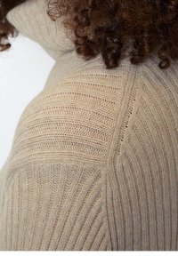 Pull en tricot beige avec texture côtelée, présentant des motifs de rayures horizontales et verticales, un col montant et une coupe ajustée.
