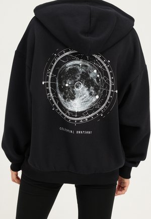 Zwarte hoodie met een grote cirkelvormige maan- en sterrenkaartgrafiek en de tekst "Celestial Rhapsody" op de rug, gedragen door een staand persoon.