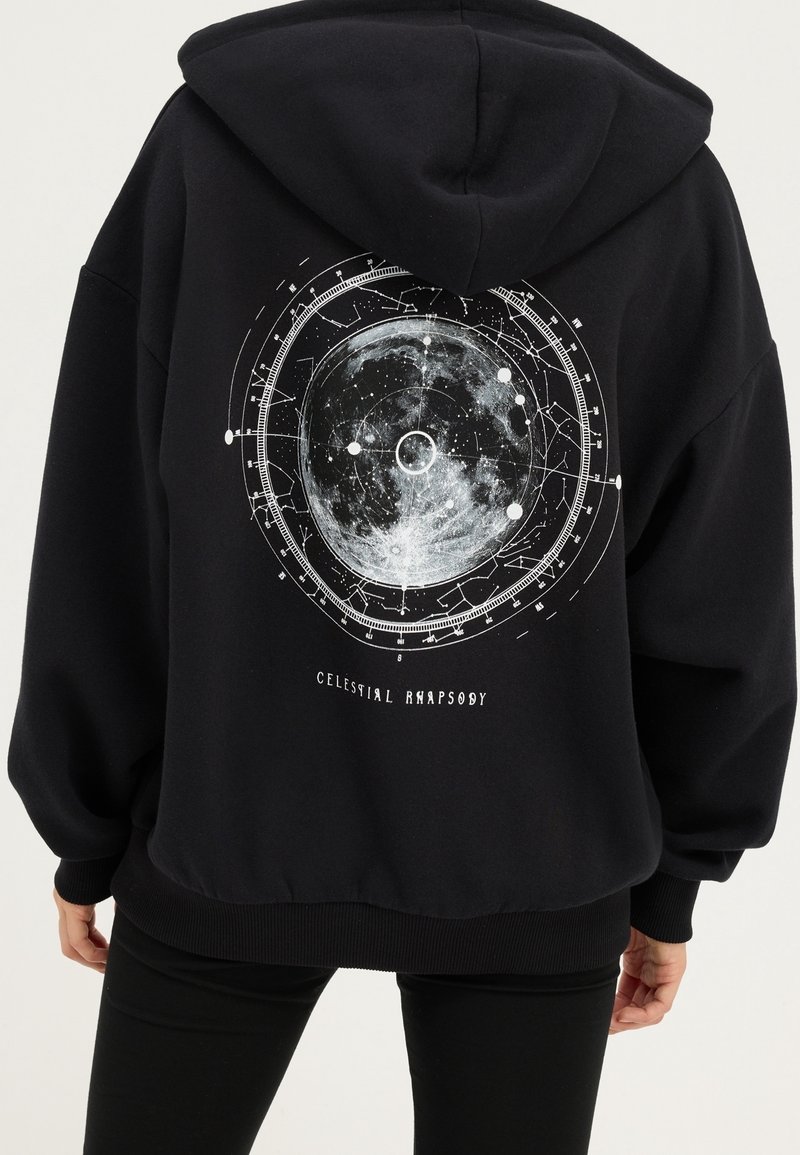 Sweat à capuche noir avec un grand graphisme circulaire de carte de la lune et des étoiles et le texte « Celestial Rhapsody » dans le dos, porté par une personne debout.