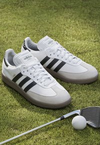 Biele kožené tenisky s šedými semišovými akcentmi, s tromi čiernymi pruhmi a gumovou podrážkou, umiestnené na tráve vedľa golfovej palice a loptičky.