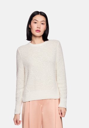 Frau mit glatten dunklen Haaren, die einen strukturierten cremefarbenen Pullover und hellrosa weit geschnittene Hosen trägt, stehend vor einem schlichten, hellen Hintergrund.