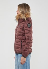 Chaqueta acolchada marrón con capucha forrada de piel sintética, diseño acolchado, bolsillos laterales y un parche de logo negro en la manga izquierda.