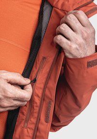 Rode buitenjas met een ritszak, mesh voering en klittenbandaccenten. Close-up van handen die de rits aanpassen.