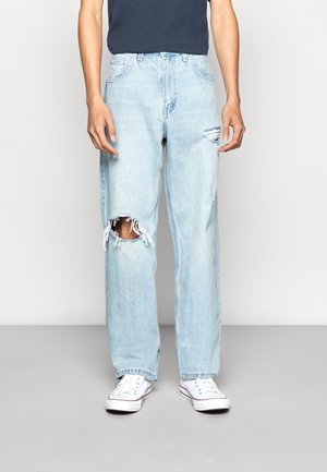 Person iført lyseblå, revnede jeans med et stort hul på venstre knæ og et mindre hul på højre lår, kombineret med hvide sneakers og en marineblå skjorte.