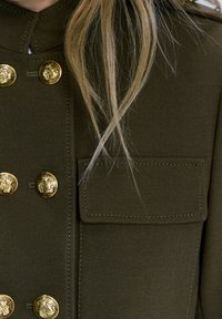 Veste militaire vert olive avec des boutons en forme de tête de lion dorés et une poche poitrine avec des détails de couture visibles. Tissu texturé.