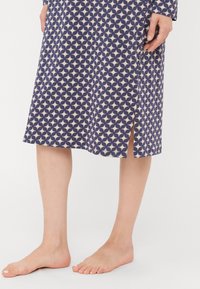 Kvinna iförd en knälång blå klänning med ett beige geometriskt mönster och en sidöppning, stående barfota mot en vit bakgrund.