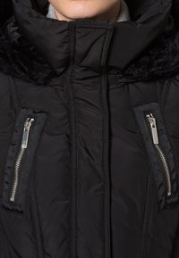 Giubbotto piumino nero con colletto alto e rifiniture in pelliccia sintetica, dotato di due tasche laterali con zip e motivo trapuntato. Texture liscia e opaca.