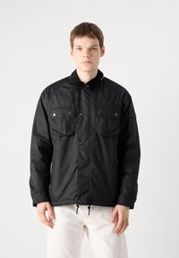 Black utility jacket with a standing collar, front snap buttons, two chest pockets, and drawstring hem. Made of durable, textured fabric.
