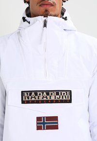 Chaqueta tipo pulóver blanca con un cuello alto, que presenta un gran parche negro y beige con el texto "NAPAPIRJI geographic" y un emblema de la bandera noruega.