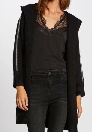 Frau trägt schwarzes Spitzen-Camisole, schwarzen Kapuzen-Cardigan mit silbernen Streifen an den Ärmeln und schwarze hoch taillierte Jeans.