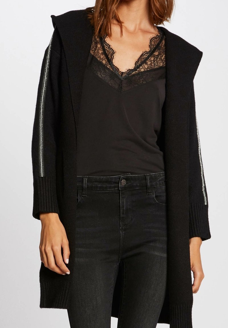 Femme portant un caraco noir bordé de dentelle, un cardigan à capuche noir avec des rayures argentées sur les manches, et un jean noir taille haute.