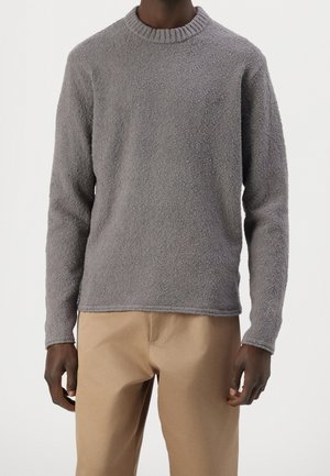 Uomo che indossa un maglione grigio a girocollo a maniche lunghe e testurizzato e pantaloni beige, in piedi contro uno sfondo semplice.