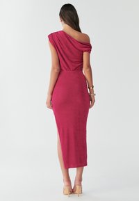 Asimetrična haljina u uskom kroju, u dubokoj boji maline. Glatka tekstura s bočnim prorezom i nabrani detalj na ramenu.