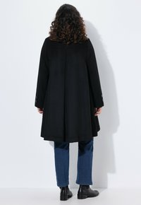 Abrigo negro de corte holgado, con mangas largas y detalles de botón en los puños. Llevado sobre jeans azules y botines. Textura suave, hasta la rodilla.