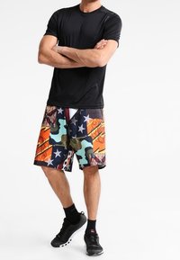 Svart kortärmad skjorta i kombination med färgglada, mönstrade shorts med stjärnor, kamouflage och abstrakta mönster. Bärs med svarta träningsskor.