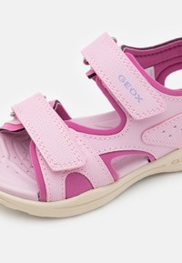 Vaaleanpunaiset lasten avonaiset sandaalit, joissa on säädettävät Velcro-nauhat ja beige pohja. Yläremmissä on "GEOX"-brändäys.