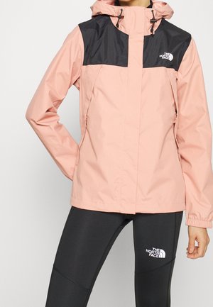Personne portant une veste à capuche North Face bicolore rose et noire et des leggings noirs North Face, debout avec une main sur la hanche.