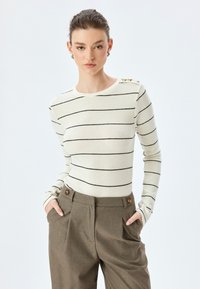 Sweter w paski w kolorze kremowym z czarnymi poziomymi liniami, posiadający długie rękawy i okrągły dekolt, ozdobiony dekoracyjnymi guzikami na ramieniu.