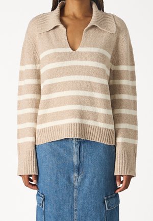 Pruhovaný béžovo-bílý pletený svetr se širokým límcem a výstřihem do V, nošený přes modrou džínovou sukni s bočními kapsami.