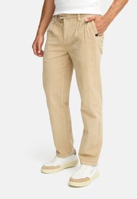 Beige corduroy trousers with pleats, front pocket, and a small logo detail. Worn with white sneakers featuring beige accents.
