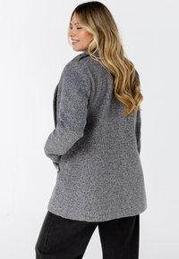 Blonde vrouw met lang golvend haar, gekleed in een gestructureerde grijze jas en zwarte broek, staande iets naar de zijkant gekeerd op een witte achtergrond.