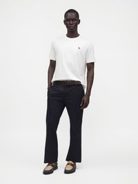 Homme debout, portant un t-shirt blanc à manches courtes, un pantalon foncé avec une ceinture marron et des mocassins marron bicolores.