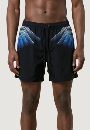 Uomo che indossa pantaloncini da nuoto neri con disegni di ali blu e grigie ai lati, in piedi con le mani rilassate ai fianchi.