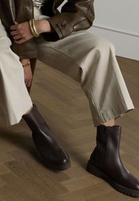 Stivaletti alla caviglia in pelle marrone con cerniera laterale, suola chunky e texture liscia, abbinati a pantaloni a gamba larga beige chiaro e una giacca marrone.