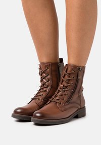 Botas de cuero marrón con cordones hasta el tobillo y cremalleras laterales, usadas sobre piernas desnudas, presentadas sobre un fondo blanco.