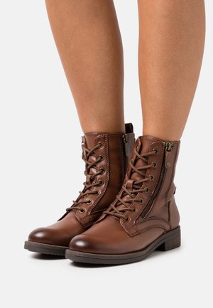 Botas de cuero marrón con cordones hasta el tobillo y cremalleras laterales, usadas sobre piernas desnudas, presentadas sobre un fondo blanco.