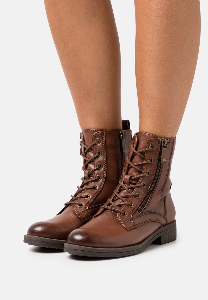 Botas de cuero marrón con cordones hasta el tobillo y cremalleras laterales, usadas sobre piernas desnudas, presentadas sobre un fondo blanco.