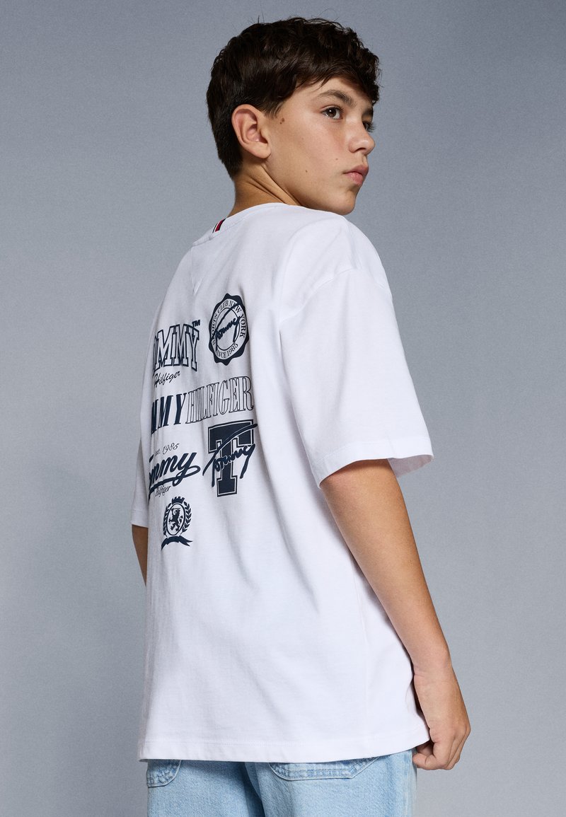 T-shirt bianco in cotone con vari loghi e design testuali neri sulla schiena, maniche corte e vestibilità rilassata. Sono indossati shorts di denim.