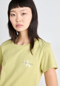 Šviesiai žalia medvilninė marškinė su apvalia iškirpte, trumpomis rankovėmis ir balta "Calvin Klein Jeans" logotipu kairėje krūtinės pusėje.