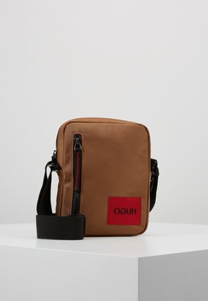 Sac bandoulière rectangulaire marron avec fermeture éclair et sangle noires, patch logo Hugo rouge à l'avant, présenté sur un socle blanc sur fond gris.