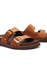 Sandales plates en cuir marron avec doubles boucles réglables et clous dorés sur une semelle rembourrée.